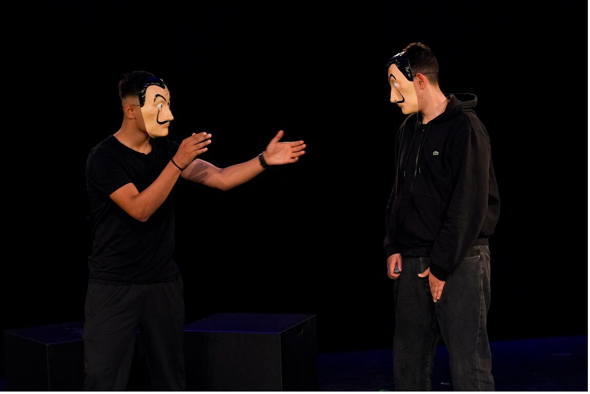 In einem schwarzen Theaterraum stehen zwei junge Männer in schwarzen Jeans, T-Shirt und Pullover, jeder mit der gleichen Plastikmaske eines männlichen Gesichts mit langem dünnen schwarzen Schnurrbart und hoch erhobenen Augenbrauen. Sie sind einander zugewandt und der eine hat seine Hände gestikulierend in Richtung des anderen erhoben.