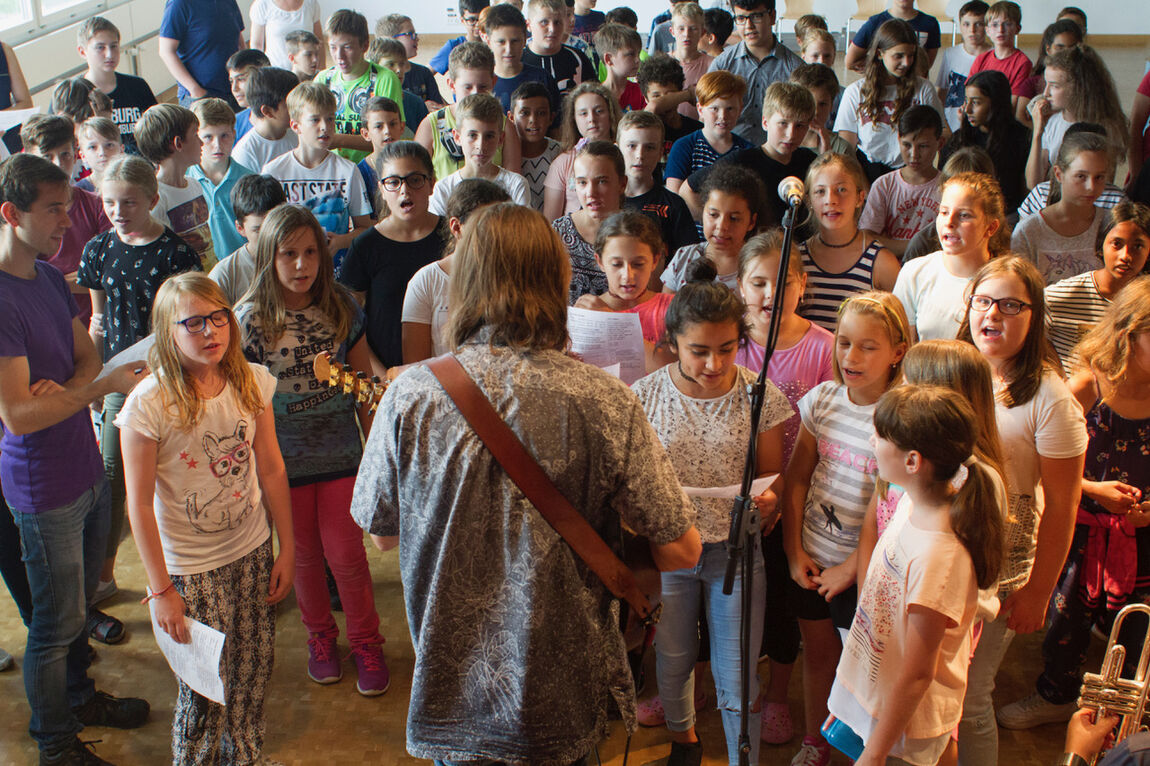 Eine grosse Gruppe von Schulkindern der Mittelstufe steht gegenüber einem Musiker mit langem grauen Haar und mit Gitarre und singt gemeinsam.