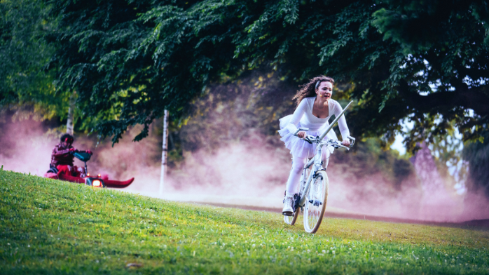 Sullo sfondo di alberi e polvere rosa che si alza dal suolo, una donna in bicicletta con un tutù bianco corre lungo un pendio erboso. Alla sua sinistra, in cima al pendio, la segue una figura vestita di viola, seduta su una slitta.