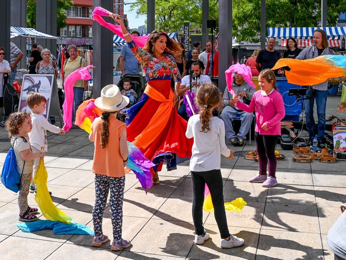 Auf einem belebten Platz tanzt eine Frau in buntem fliegenden Rock und schwingt ein Tuch. Kinder stehen um sie herum und schwingen ebenfalls bunte Tücher. Ein Gitarrist begleitet sie.