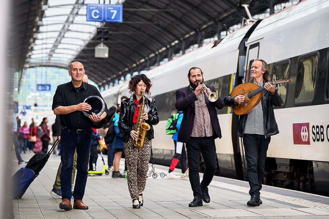 vier Musiker_Innen gehen nebeneinander auf dem Perron eines Bahnhofs musizierend entlang mit Trommel, Saxophon, Trompete und Oud. 