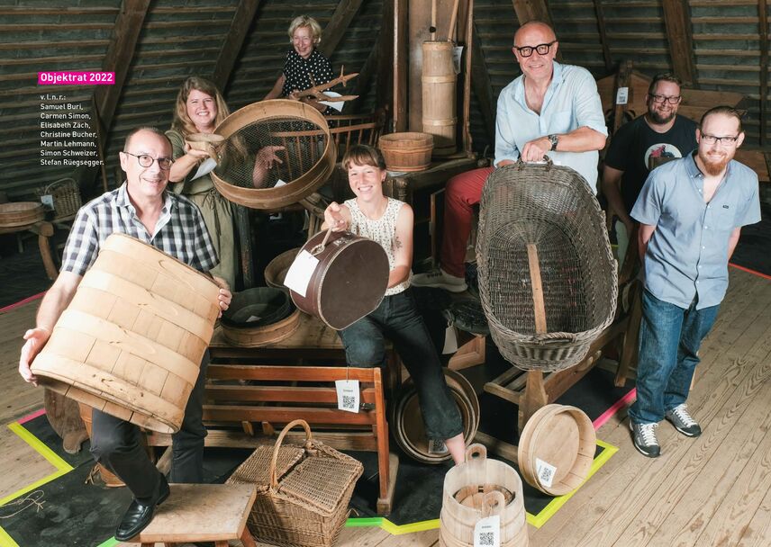 Auf dem Dachstock eines alten Gebäudes aus Holz posieren vier Männer und drei Frauen mit alten Körben, Koffern und Gefässen aus Holz und lachen in dien Kamera.